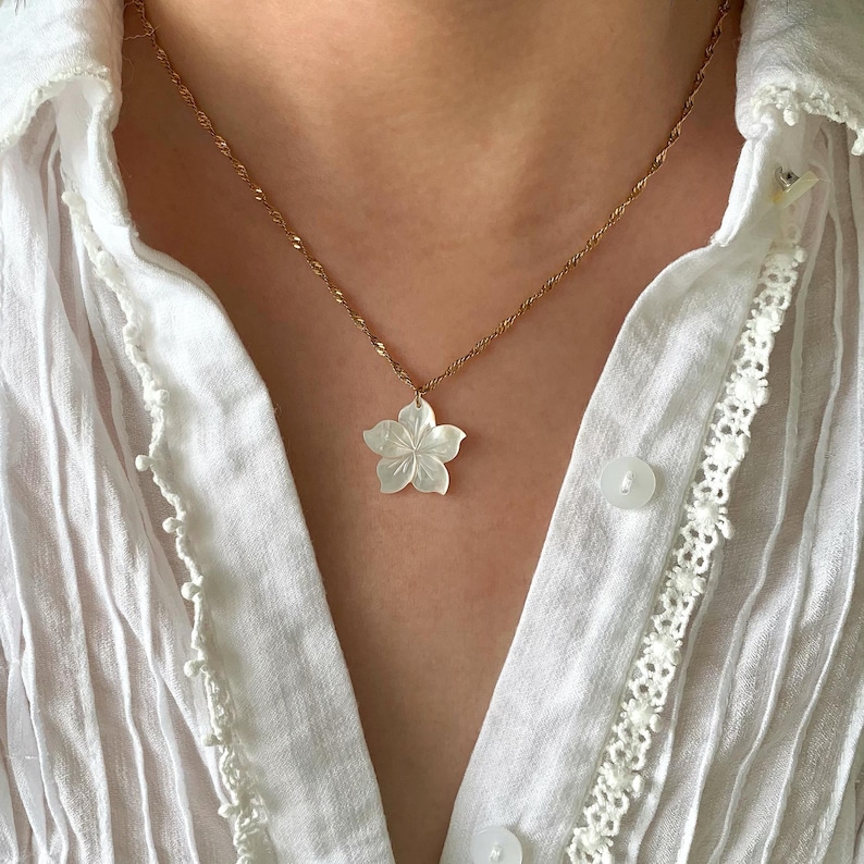 May include: A gold-colored chain necklace with a white, flower-shaped pendant. The pendant has a detailed, carved design. The necklace is worn over a white, button-down shirt.