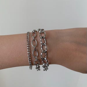 May include: Three silver chain bracelets stacked on a wrist. The bracelets are made of different chain styles, including a curb chain, a rope chain, and a link chain.
