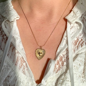 Puede incluir: Un collar con colgante de corazón dorado. El colgante tiene un diseño texturizado de rayos de sol con una pequeña piedra transparente y redonda en el centro. El collar se lleva sobre una blusa de encaje blanco.