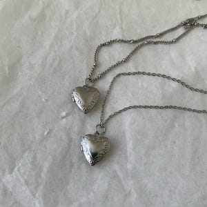 以下が含まれることがあります： 複雑なデザインが施された、銀色のハート型のロケットネックレスが2つ。ロケットは開いており、チェーンは互いにつながっています。