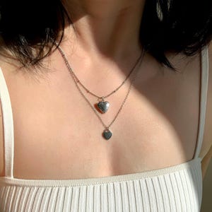 以下が含まれることがあります： 2つのハート型のペンダントが付いたシルバーの重ね付けネックレス。上のペンダントは、細かなデザインが施されたロケットで、下のペンダントはシンプルなハートです。
