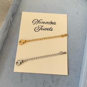May include: Two necklace extenders, one gold-toned and one silver-toned, are displayed on a cream-coloured card. The card has the text "November Jewels" in elegant script. The extenders are on a light blue surface.