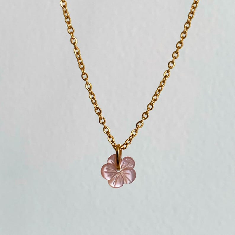 May include: A delicate gold chain necklace with a pink flower pendant. The flower is carved from a translucent material and has a delicate, feminine design.