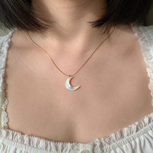 May include: A gold chain necklace featuring a white, pearlescent crescent moon pendant. The necklace is worn around the neck. The pendant is a delicate piece of jewellery.