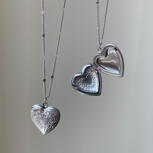 May include: Two silver heart-shaped locket necklaces with intricate designs. The lockets are open, revealing the inside of the heart.