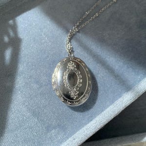 May include: A silver-colored oval locket necklace with an ornate design. The locket hangs from a delicate chain and is displayed on a blue surface. The locket features a detailed, symmetrical pattern around the edges and center.