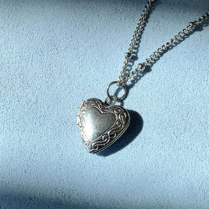May include: A silver-toned heart-shaped locket pendant suspended from a delicate metal chain. The locket features an intricate embossed scrollwork pattern along its outer border, with a smooth, polished center surface and a small circular jump ring.