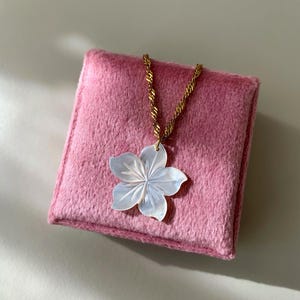 May include: A gold chain necklace with a white flower pendant, resting on a pink, square, plush surface. The flower pendant has detailed petal carvings. The necklace is displayed on a light background.