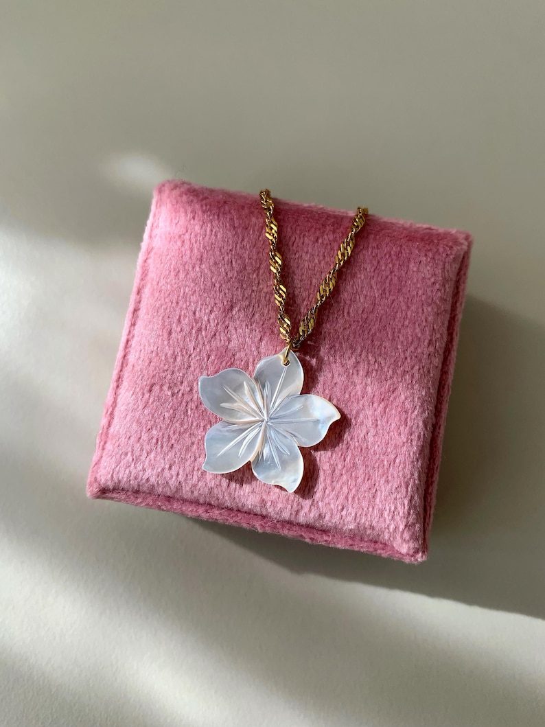 May include: A gold chain necklace with a white flower pendant, resting on a pink, square, plush surface. The flower pendant has detailed petal carvings. The necklace is displayed on a light background.