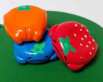 May include: Three colorful fruit-shaped figurines: an orange, a blue blueberry, and a red strawberry. Each has a smiling face and green accents. They are arranged on a green, leaf-shaped surface, creating a playful and whimsical scene.