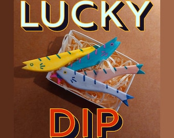 Puede incluir: Una caja blanca llena de virutas de madera y tres peces de madera. Los peces están pintados de amarillo, azul y rosa. La caja tiene las palabras "LUCKY DIP" impresas en la parte delantera.