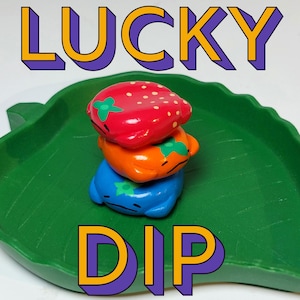 Puede incluir: Un plato verde en forma de hoja contiene una pila de tres figuras coloridas con forma de fruta de cerámica. La figura superior es roja con puntos blancos, la del medio es naranja y la inferior es azul. La palabra "LUCKY" está arriba y "DIP" abajo.
