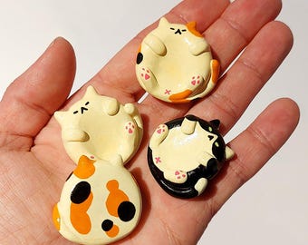 May include: Three small, round, ceramic-like cat figurines in a palm. One is cream with orange and black patches, one is solid cream with orange accents, and one is black and white. Each cat is depicted lying on its back with visible pink paw pads.