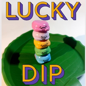 May include: A stack of five colorful, rounded figures with sad faces, in pink, lavender, yellow, green, and blue, sits on a green leaf-shaped dish. The word "LUCKY" is above the figures, and "DIP" is below, both in a 3D font.