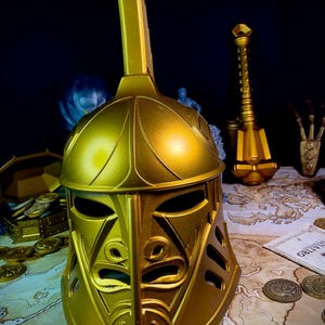 K&ouml;nnte beinhalten: Ein goldfarbener Helm mit einem hohen, rechteckigen Oberteil und einem detaillierten Gesichtsdesign. Der Helm ist Teil einer Sammlung von Fantasy-Artikeln, darunter ein Streitkolben, M&uuml;nzen und eine Schatztruhe, die alle auf einer Karte ausgestellt sind.