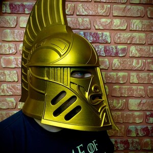 K&ouml;nnte beinhalten: Ein goldfarbener Helm mit hohem, geriffeltem Kamm und aufwendigen Gesichtsdetails. Der Helm hat Augenschlitze und ein stilisiertes Gesichtsdesign. Die Person, die den Helm tr&auml;gt, tr&auml;gt ein schwarzes T-Shirt mit den Worten "GAME OF".