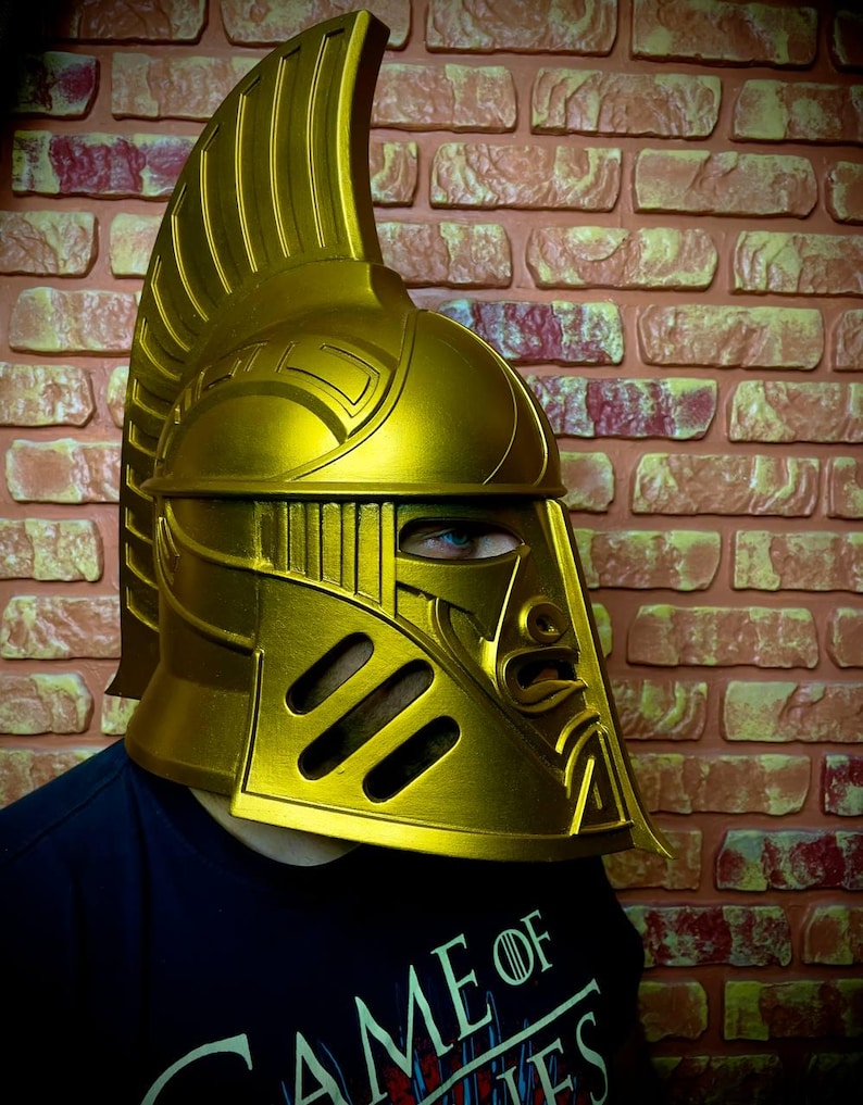K&ouml;nnte beinhalten: Ein goldfarbener Helm mit hohem, geriffeltem Kamm und aufwendigen Gesichtsdetails. Der Helm hat Augenschlitze und ein stilisiertes Gesichtsdesign. Die Person, die den Helm tr&auml;gt, tr&auml;gt ein schwarzes T-Shirt mit den Worten "GAME OF".