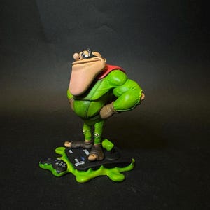 K&ouml;nnte beinhalten: Figur einer Zeichentrickfigur in einem gr&uuml;nen Outfit mit rotem Umhang, braunen Stiefeln und einer gro&szlig;en Nase. Die Figur steht auf einer gr&uuml;nen, bespritzten Basis mit einem Gamecontroller.