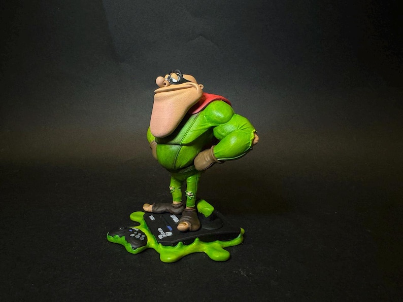 K&ouml;nnte beinhalten: Figur einer Zeichentrickfigur in einem gr&uuml;nen Outfit mit rotem Umhang, braunen Stiefeln und einer gro&szlig;en Nase. Die Figur steht auf einer gr&uuml;nen, bespritzten Basis mit einem Gamecontroller.