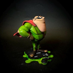 K&ouml;nnte beinhalten: Figur eines Comic-Charakters in einem gr&uuml;nen Overall und rotem Umhang, der eine schwarze Maske tr&auml;gt. Die Figur steht auf einer schwarzen Basis mit gr&uuml;nem Schleim und dem Text "MEGA 32 BIT".