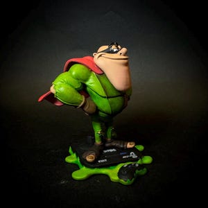 K&ouml;nnte beinhalten: Figur eines Comic-Charakters in einem gr&uuml;nen Overall und rotem Umhang, der eine schwarze Maske tr&auml;gt. Die Figur steht auf einer schwarzen Basis mit gr&uuml;nem Schleim und dem Text "MEGA DRIVE" und "16BIT".