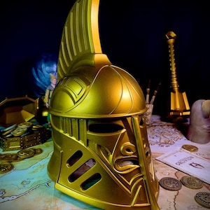 K&ouml;nnte beinhalten: Ein goldfarbener Helm mit hohem, vertikalem Kamm und aufwendigen Details. Der Helm hat Augen- und Mund&ouml;ffnungen und wird auf einer Karte mit M&uuml;nzen und einer Schatztruhe ausgestellt. Das Gesamtthema ist Fantasy oder Historie.