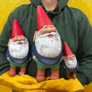 Puede incluir: Tres gnomos de jardín de diferentes tamaños. Cada gnomo tiene un gorro puntiagudo rojo, una barba blanca y una cara sonriente. Visten chaquetas azules, pantalones verdes y botas marrones. Los gnomos se sostienen sobre un fondo amarillo.