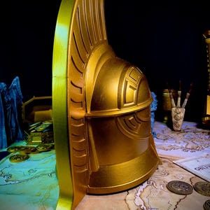 K&ouml;nnte beinhalten: Ein goldfarbener Helm mit detailliertem Design, mit einem hohen, geriffelten Kamm und komplizierten Mustern. Der Helm wird auf einer Karte mit M&uuml;nzen und anderen Requisiten pr&auml;sentiert, wodurch ein Fantasy-Thema entsteht. Die metallische Oberfl&auml;che reflektiert das Licht.