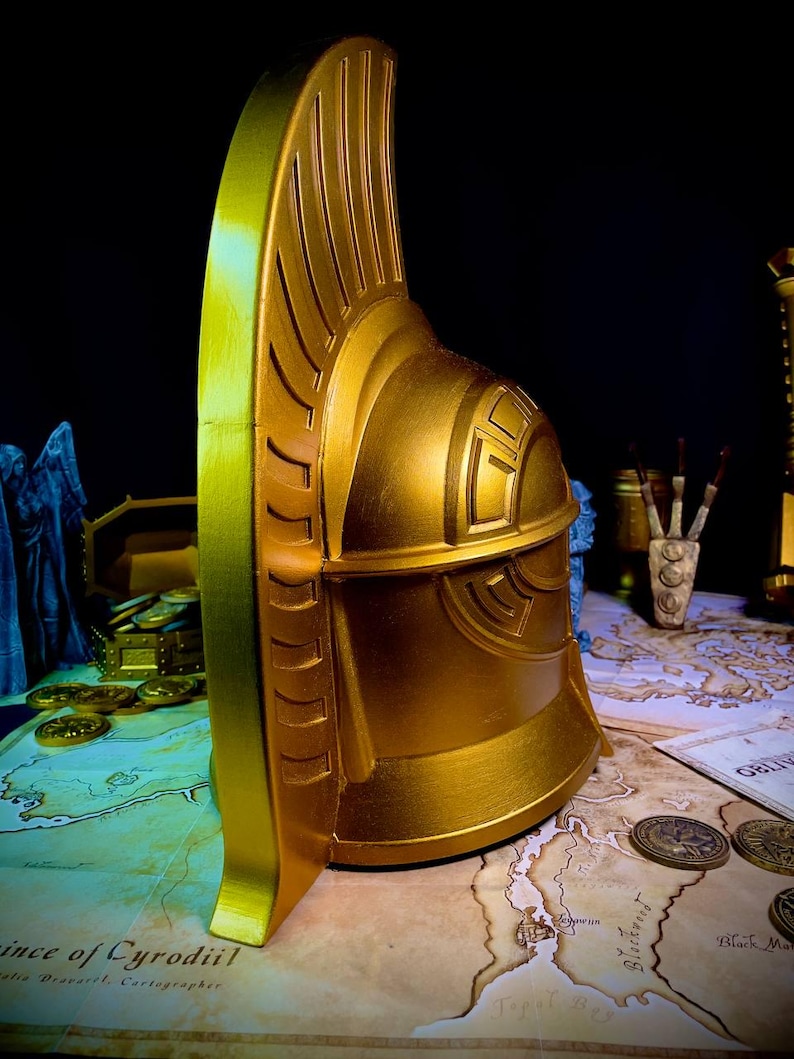 K&ouml;nnte beinhalten: Ein goldfarbener Helm mit detailliertem Design, mit einem hohen, geriffelten Kamm und komplizierten Mustern. Der Helm wird auf einer Karte mit M&uuml;nzen und anderen Requisiten pr&auml;sentiert, wodurch ein Fantasy-Thema entsteht. Die metallische Oberfl&auml;che reflektiert das Licht.