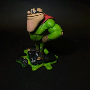 K&ouml;nnte beinhalten: Detaillierte Figur einer Zeichentrickfigur in einem gr&uuml;nen Outfit mit rotem Umhang und schwarzer Brille. Die Figur steht auf einer schwarzen Plattform mit gr&uuml;nem, gesprenkeltem Sockel. Die Figur hat eine gro&szlig;e Nase und einen selbstbewussten Ausdruck.