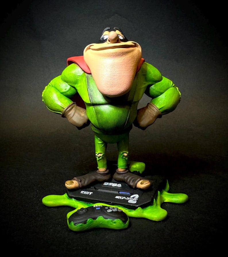 K&ouml;nnte beinhalten: Eine gr&uuml;ne Figur einer Zeichentrickfigur mit gro&szlig;em Kinn, rotem Umhang und braunen Handschuhen. Die Figur steht auf einer schwarzen Sega Mega Drive Konsole mit einem gr&uuml;nen Controller und einer gr&uuml;nen, klebrigen Basis. Das Mega Drive Logo ist sichtbar.