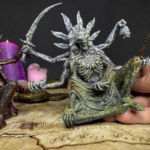 Op de afbeelding: Een gedetailleerd, handgeschilderd beeldje van een skeletgodheid met meerdere armen, die een gebogen mes en een afgehakt hoofd vasthoudt. De figuur zit, met een groen-grijze kleurstelling. Een tweede beeldje is zichtbaar.