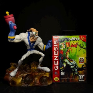Pode incluir: Estatueta detalhada de Earthworm Jim em um traje branco e azul, segurando uma arma vermelha e amarela. Ao lado, uma caixa de jogo Sega Genesis com o título e a arte do personagem Earthworm Jim. Fundo preto.