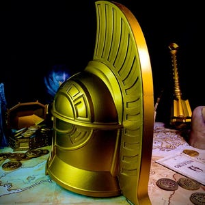 K&ouml;nnte beinhalten: Ein goldener Helm mit hohem, verziertem Kamm vor dunklem Hintergrund. Der Helm weist detaillierte Rillen und geometrische Muster auf. Eine Karte und M&uuml;nzen im Hintergrund deuten auf ein Fantasy- oder historisches Thema hin.