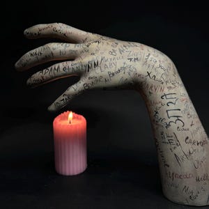Könnte beinhalten: Eine Gipshandskulptur, die mit Schrift bedeckt ist und eine brennende rosa Kerze hält. Die Schrift umfasst Namen, Daten und andere Botschaften.