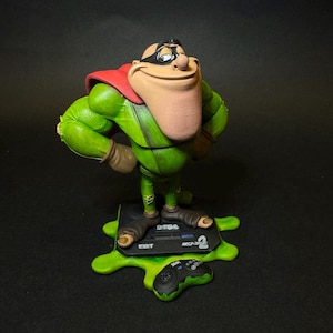 K&ouml;nnte beinhalten: Gr&uuml;ne Figur einer Zeichentrickfigur mit rotem Umhang und schwarzer Maske, die die H&auml;nde in die H&uuml;ften gestemmt hat. Die Figur steht auf einem Sockel mit einer Sega Mega Drive Konsole und Controller, alles auf einer gr&uuml;nen, strukturierten Oberfl&auml;che. Der Sockel tr&auml;gt die Aufschrift "16BIT".