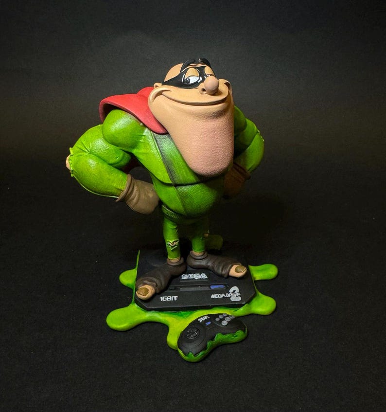 K&ouml;nnte beinhalten: Gr&uuml;ne Figur einer Zeichentrickfigur mit rotem Umhang und schwarzer Maske, die die H&auml;nde in die H&uuml;ften gestemmt hat. Die Figur steht auf einem Sockel mit einer Sega Mega Drive Konsole und Controller, alles auf einer gr&uuml;nen, strukturierten Oberfl&auml;che. Der Sockel tr&auml;gt die Aufschrift "16BIT".