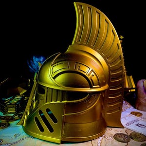 K&ouml;nnte beinhalten: Ein goldfarbener Helm mit hohem, stilisiertem Kamm und komplizierten geometrischen Mustern. Der Helm hat ein Visier und Wangenschutz. Er wird auf einer Karte mit verstreuten M&uuml;nzen ausgestellt, was auf ein Fantasy- oder historisches Thema hindeutet.