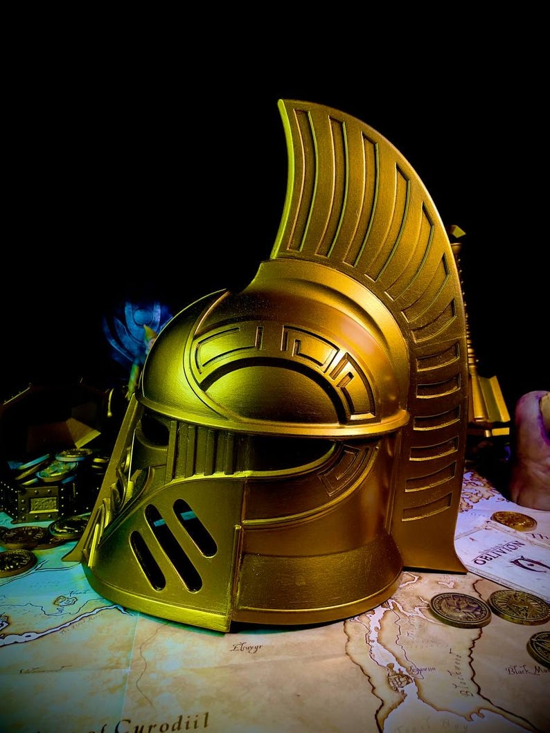 K&ouml;nnte beinhalten: Ein goldfarbener Helm mit hohem, stilisiertem Kamm und komplizierten geometrischen Mustern. Der Helm hat ein Visier und Wangenschutz. Er wird auf einer Karte mit verstreuten M&uuml;nzen ausgestellt, was auf ein Fantasy- oder historisches Thema hindeutet.