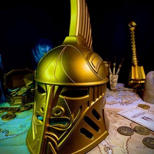 K&ouml;nnte beinhalten: Ein goldfarbener Helm mit aufwendigen Details und einem hohen, vertikalen Kamm. Der Helm hat einen Gesichtsschutz mit einem stilisierten Design. Der Hintergrund zeigt eine Karte, M&uuml;nzen und einen Streitkolben, was auf ein Fantasy- oder historisches Thema hindeutet.