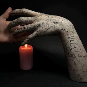 Könnte beinhalten: Eine Gipshand, die mit Schrift bedeckt ist, greift nach einer brennenden rosa Kerze. Die Schrift umfasst Namen, Daten und andere Botschaften.