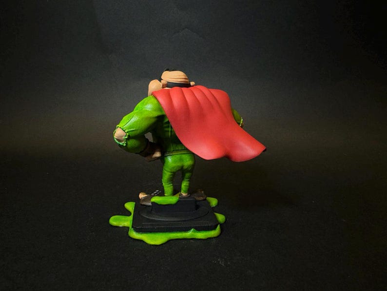 K&ouml;nnte beinhalten: Figur eines Charakters in gr&uuml;nem Outfit und rotem Umhang, stehend auf einer schwarzen Basis mit gr&uuml;nem Schleim. Die Figur hat einen beigen Hut und posiert mit den Armen an den Seiten. Der Hintergrund ist dunkel.