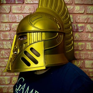 K&ouml;nnte beinhalten: Ein goldfarbener Helm mit hohem, geriffeltem Kamm und aufwendigen Details. Der Helm hat einen Gesichtsschutz mit Augenschlitzen und ein stilisiertes Design. Die Person tr&auml;gt ein dunkelblaues T-Shirt mit der Aufschrift "GAME OF".