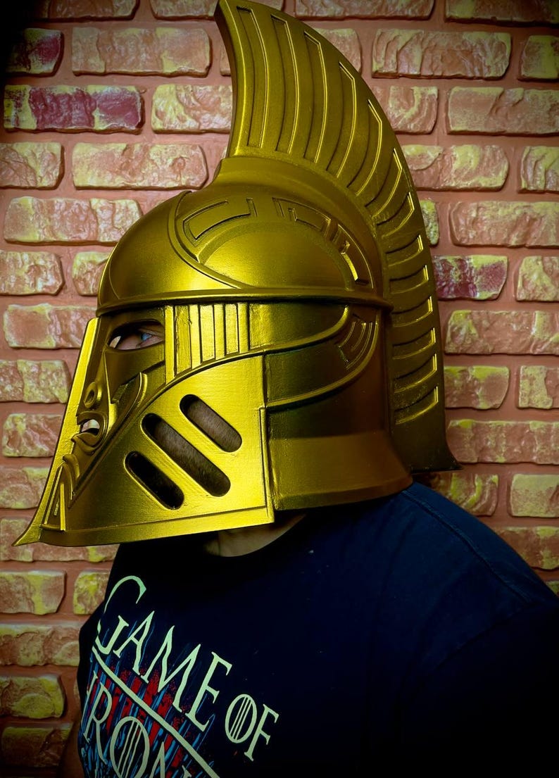 K&ouml;nnte beinhalten: Ein goldfarbener Helm mit hohem, geriffeltem Kamm und aufwendigen Details. Der Helm hat einen Gesichtsschutz mit Augenschlitzen und ein stilisiertes Design. Die Person tr&auml;gt ein dunkelblaues T-Shirt mit der Aufschrift "GAME OF".