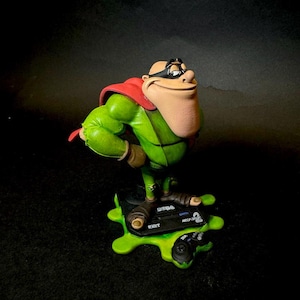 K&ouml;nnte beinhalten: Eine Figur einer gr&uuml;nen Figur mit rotem Umhang und schwarzer Maske, die auf einer gr&uuml;nen Basis mit einem Controller und dem Text "SEGA 16BIT ARCADE" steht. Die Figur hat ein gro&szlig;es Kinn und posiert mit einem erhobenen Arm.