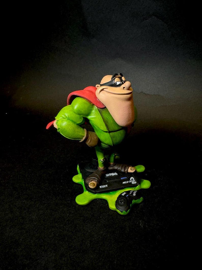 K&ouml;nnte beinhalten: Eine Figur einer gr&uuml;nen Figur mit rotem Umhang und schwarzer Maske, die auf einer gr&uuml;nen Basis mit einem Controller und dem Text "SEGA 16BIT ARCADE" steht. Die Figur hat ein gro&szlig;es Kinn und posiert mit einem erhobenen Arm.