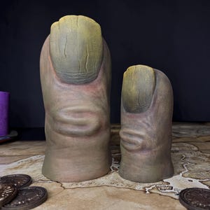 May include: Two ceramic finger sculptures with detailed skin texture and large, yellow-green nails. The fingers are a fleshy tone, and the sculptures are displayed on a map with bronze coins.