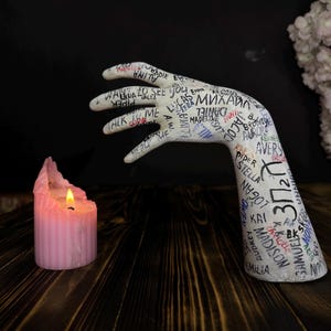 Peut inclure: Sculpture de main en céramique blanche avec une surface texturée. La main est recouverte de noms et de mots écrits à la main, notamment "Talk to Me", "See You" et "312".