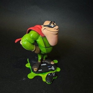 K&ouml;nnte beinhalten: Figur einer Zeichentrickfigur in gr&uuml;nem Outfit, rotem Umhang und schwarzer Maske. Die Figur steht auf einer gr&uuml;nen Basis mit einem Videospiel-Controller und dem Text "SEGA MEGA DRIVE".