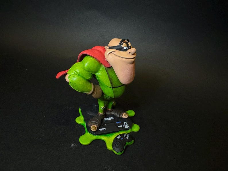 K&ouml;nnte beinhalten: Figur einer Zeichentrickfigur in gr&uuml;nem Outfit, rotem Umhang und schwarzer Maske. Die Figur steht auf einer gr&uuml;nen Basis mit einem Videospiel-Controller und dem Text "SEGA MEGA DRIVE".